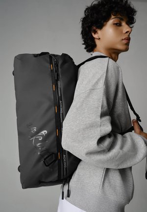 Sac à dos gris avec des accents noirs, doté d'une fermeture éclair, d'une surface texturée et d'un détail de logo. Porté sur le dos d'un mannequin par-dessus un sweatshirt gris.