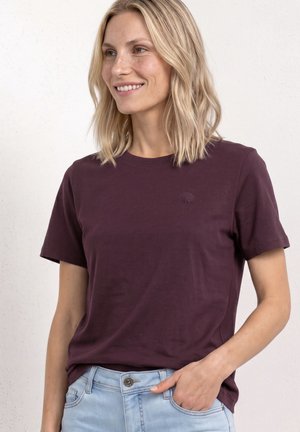 Blonde vrouw draagt een effen donkerpaarse t-shirt en lichtblauwe spijkerbroek, glimlacht en kijkt iets naar links met één hand in haar zak.