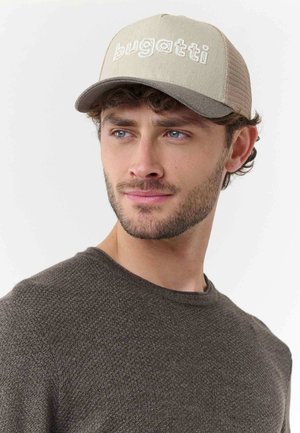 Young man with short curly hair and beard wearing a beige Bugatti cap and dark textured sweater, looking slightly to his right.