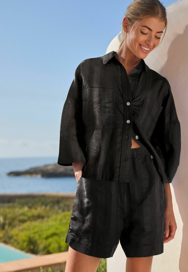 Sort linned to-delt outfit bestående af en knap-op skjorte med krave og påsyede lommer, parret med matchende shorts. Struktureret stof med striber.