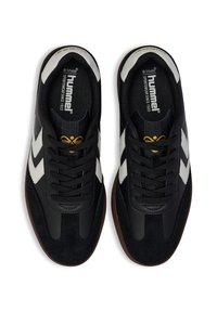 Par tenisica crne boje marke Hummel s bijelim chevron prugama, smešnim potplatima, crnim vezicama i zlatnim logotipom na jeziku, prikazane odozgo na bijeloj pozadini.