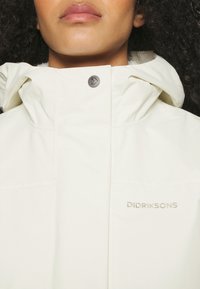 Chaqueta impermeable blanca con cuello alto, cierre de cremallera, dos bolsillos frontales y un logo bordado en dorado. Detalle de botón a presión plateado.