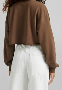 Felpa marrone corta con maniche lunghe, realizzata in materiale morbido, abbinata a pantaloni bianchi a vita alta con tasche e una silhouette aderente.