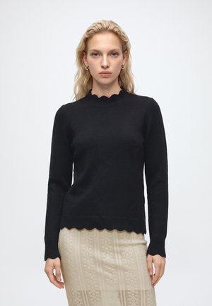Femme blonde portant des boucles d'oreilles créoles dorées, un pull noir à manches longues avec des bords festonnés et une jupe beige à motifs, faisant face vers l'avant.