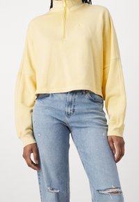 Keltainen lyhyt collegepaita, jossa on korkea kaulus ja neljänneksen vetoketju. Siinä on hillitty logo ja rennot hihat. Yhdistetty kuluneisiin sinisiin farkkuihin.