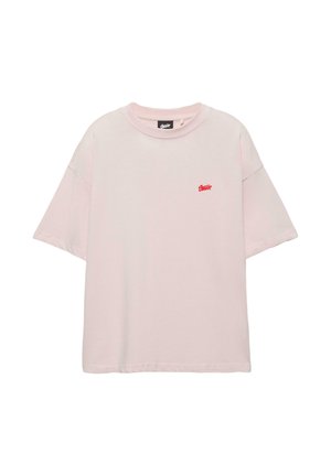 Tricou roz deschis, supradimensionat, cu mâneci scurte, cu un mic logo roșu pe pieptul stâng și decolteu rotund.