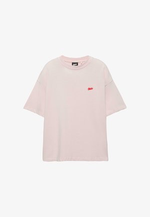 Jasnoróżowa, luźna koszulka z krótkim rękawem, z małym czerwonym logo na lewej piersi i okrągłym dekoltem.