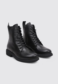 Bottes chelsea en cuir noir à lacets, avec un bout rond et des semelles plates. Texture lisse avec un design minimaliste et des fermetures éclair latérales.