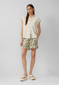 Chemise boutonnée beige clair à manches courtes, associée à un short vert olive avec des motifs floraux. Modèle portant des sandales à plateforme légères.