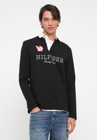 Melna garām piedurknēm polo krekls ar baltu apkakli, rotāts ar izšūtu "HILFIGER Racing Club" tekstu un sarkano logo uzlīmi kreisajā krūtī.