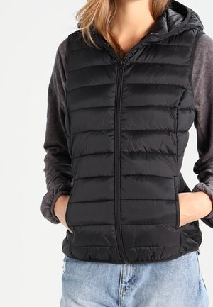 Vrouw met een zwarte gewatteerde bodywarmer over een grijze lange mouwtop en blauwe jeans, met de handen in de zakken van het vest, getoond van schouders tot heupen.