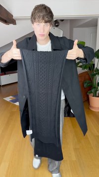 Czarny sweter dziany na drutach, z fakturowanym wzorem warkocza, długimi rękawami i prostokątnym kształtem. Materiał wydaje się gruby i ciepły.