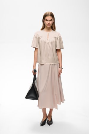 Femme aux cheveux raides portant un chemisier beige à manches courtes et une jupe longue assortie, tenant un sac à main noir, debout sur un fond blanc.