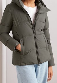 Oliivinvihreä toppatakki hupulla, sivutaskuilla ja edessä vetoketjulla. Takissa on tikattu pinta ja pehmeä kangas.