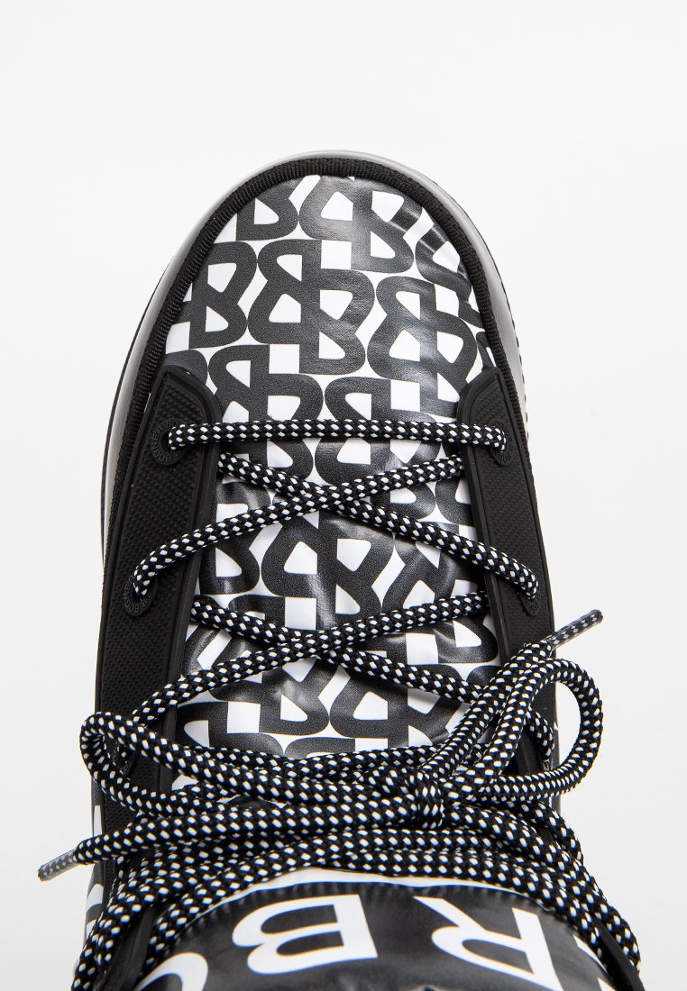 Nahaufnahme der Schuhspitze eines schwarz-weißen Sneakers mit sich wiederholendem "B"-Muster und schwarz-weißen geflochtenen Schnürsenkeln, die zu einer Schleife gebunden sind.
