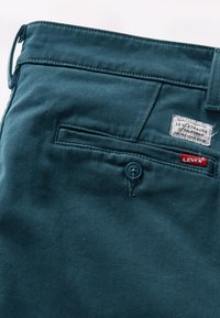 Pantaloni din denim teal, cu un buzunar la spate, buton albastru și etichetă Levi's. Textură netedă, cu cusături contrastante și un luciu subtil.