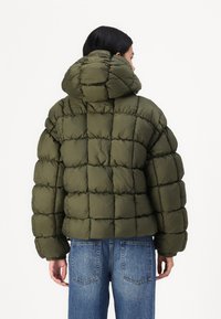 Olivenfarget pufferjakke med hette, quiltet design og elastiske mansjetter, paret med høyhårede blå jeans.
