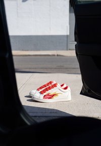 Des baskets blanches avec des lacets rouges et un motif de flammes jaune-rouge sont posées sur un trottoir en béton à côté d’une portière de voiture ouverte par une journée ensoleillée.