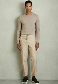 Jersey beige de manga larga combinado con pantalones beige claros que cuentan con una cintura elástica y puños. Zapatillas negras con acentos oscuros.