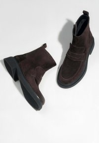 Tmav�ě hnědé semišové kotníkové boty s černou gumovou podrážkou, bočním zipem a ozdobným páskem na světlém pozadí.