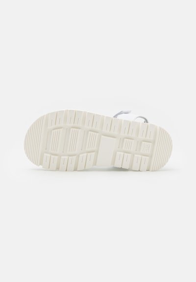 Semelle en caoutchouc blanche avec des rainures et des arêtes texturées, présentant un design plat et un bout arrondi. Profil minimaliste avec un branding subtil.