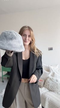 Harmaa tekoturkisesta valmistettu bucket-hattu, jota pitää henkilö musta bleiseri päällään. Taustalla näkyy pehmeitä sisustuselementtejä ja vihreä pyörillä varustettu kärry.