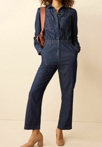 Combinaison en denim bleu foncé avec un col, fermeture à boutons, deux poches poitrine, taille élastique et jambes larges. Associée à des talons beiges.