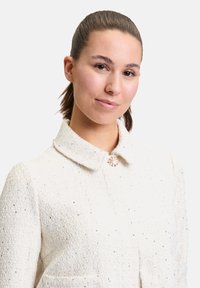 Witte textuurjasje met een kraag, voorzien van een decoratieve zilveren knoop en kleine pailletten door de stof heen. Zakjes zijn inbegrepen.