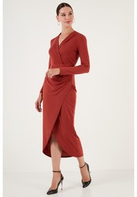 Robe longue rouge à manches longues avec une fente jusqu'au genou et des détails froncés sur le côté, fabriquée en tissu doux. Portée avec des chaussures à talons hauts noires.