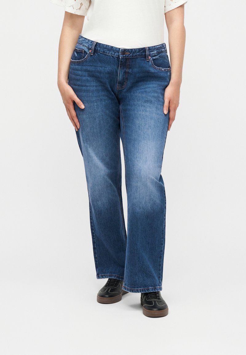 Femme portant un jean bleu droit, des baskets noires et un haut blanc avec des manches en dentelle à œillets, debout sur un fond blanc.