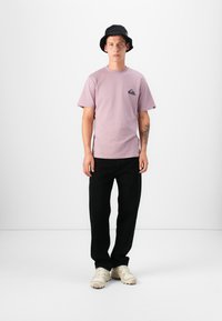 Lyse lilla kortermet t-skjorte med lomme-logo, svarte bukser og beige joggesko. Modell som bærer en svart bøttehatt. Enkel design, avslappet passform.