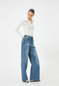 Polo a maniche lunghe bianco a coste, leggero, abbinato a jeans in denim azzurro chiaro a vita alta e gamba larga. Caratterizzato da una vestibilità rilassata e da un leggero sbiadimento.