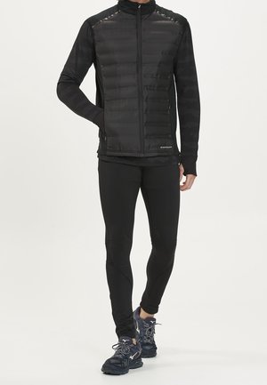 Mand iført sort polstret lynlåstrøje, sorte leggings og sorte løbesko, stående med den ene hånd i jakke lommen.