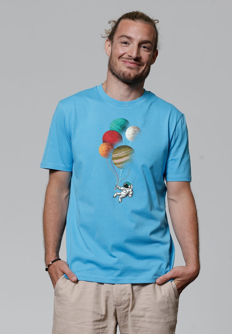T-shirt bleu clair à manches courtes mettant en avant un astronaute tenant des planètes colorées en forme de ballons, avec des graphismes vibrants et un design décontracté.