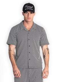 Camisa de botones de manga corta en negro con un patrón geométrico blanco; combinada con una gorra negra con texto y detalles del logo en blanco.