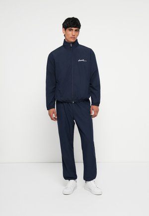 Navy windbreaker with high collar and zipper, featuring elastic cuffs and waistband. Paired with matching pants and white sneakers.