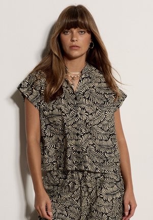 Jeune femme aux longs cheveux bruns portant un haut et un short assortis noirs et beiges à motif feuillu, debout devant un fond clair uni.