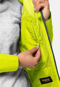 Giacca gialla brillante con fodera in rete e interno in pelliccia sintetica. Presenta una tasca con zip e un'etichetta con il logo. Dettaglio della tasca con zip a mano.