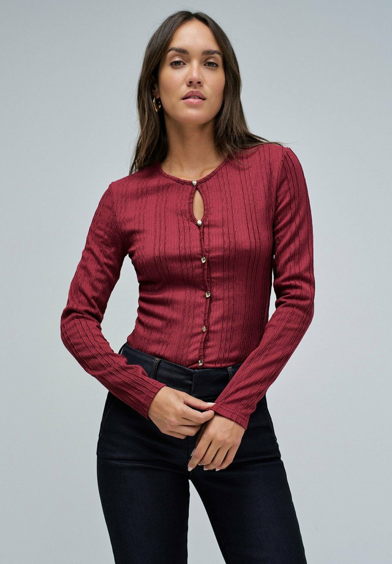 Top burdeos de manga larga con textura estriada vertical, escote tipo ojal y cierre de botón. Llevado con pantalones de mezclilla oscuros.