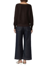 Personne portant un pull à manches longues marron foncé, un jean en denim large, et des chaussures à talon bas blanches, se tenant de dos.