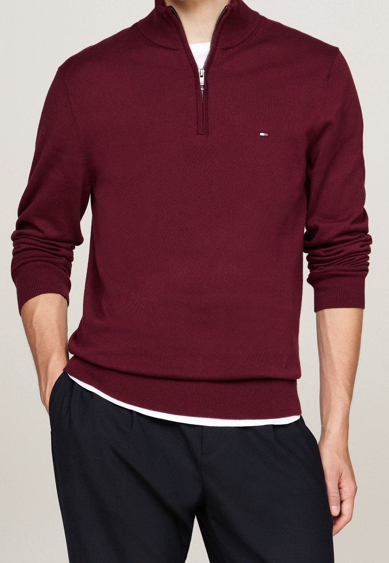 Homme portant un pull bordeaux manches longues avec fermeture éclair quart-zip sur une chemise blanche, associé à un pantalon noir, main dans la poche.