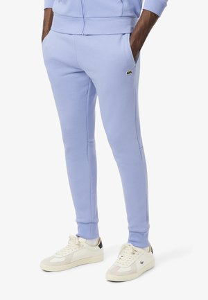 Pantalones deportivos de color púrpura claro, confeccionados en mezcla de algodón, con bolsillos laterales, costuras en contraste y un pequeño logo verde bordado. Combinados con zapatillas blancas.