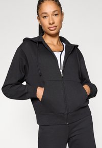 Μαύρο φούτερ με φερμουάρ, κουκούλα με κορδόνι, τσέπες καγκουρό και ριμπ μανσέτες. Κατασκευασμένο από απαλό, υφή ύφασμα.