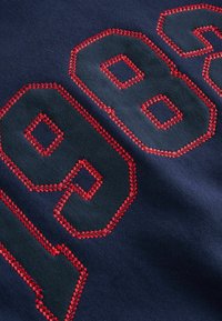 Tissu bleu marine avec chiffres brodés rouges "1986" ornés d'accents en forme de diamant sur les bords. Texture douce avec détail de couture visible.