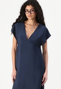 Rochie bleumarin cu mâneci scurte, de tip cap-sleeve, și decolteu adânc în V. O țesătură moale, ușor texturată, care cade liber de la talie, creând o siluetă relaxată.