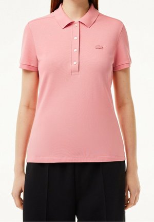 Femme portant un polo rose à manches courtes avec des boutons et un petit logo brodé, associé à un pantalon noir, sur fond blanc.