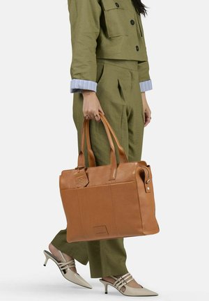 Sac tote en cuir tanné avec poignées doubles, texture lisse, poche zippée avant et logo embossé. Porté avec une tenue olive et des talons beiges.