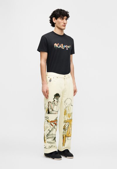 Μαύρο βαμβακερό T-shirt με πολύχρωμο γραφικό σχέδιο, συνδυασμένο με ελεφαντόδοντα τζιν με καλλιτεχνικά σκίτσα, συμπληρωμένο με μαύρα αθλητικά παπούτσια.