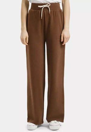 Brązowe spodnie o szerokich nogawkach z elastycznym paskiem i białym sznurkiem, noszone z białymi tenisówkami na stojącej osobie.