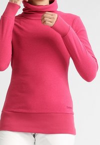 Mujer ajustando el cuello de un suéter de cuello alto rosa de manga larga, vistiendo pantalones blancos frente a un fondo gris liso.